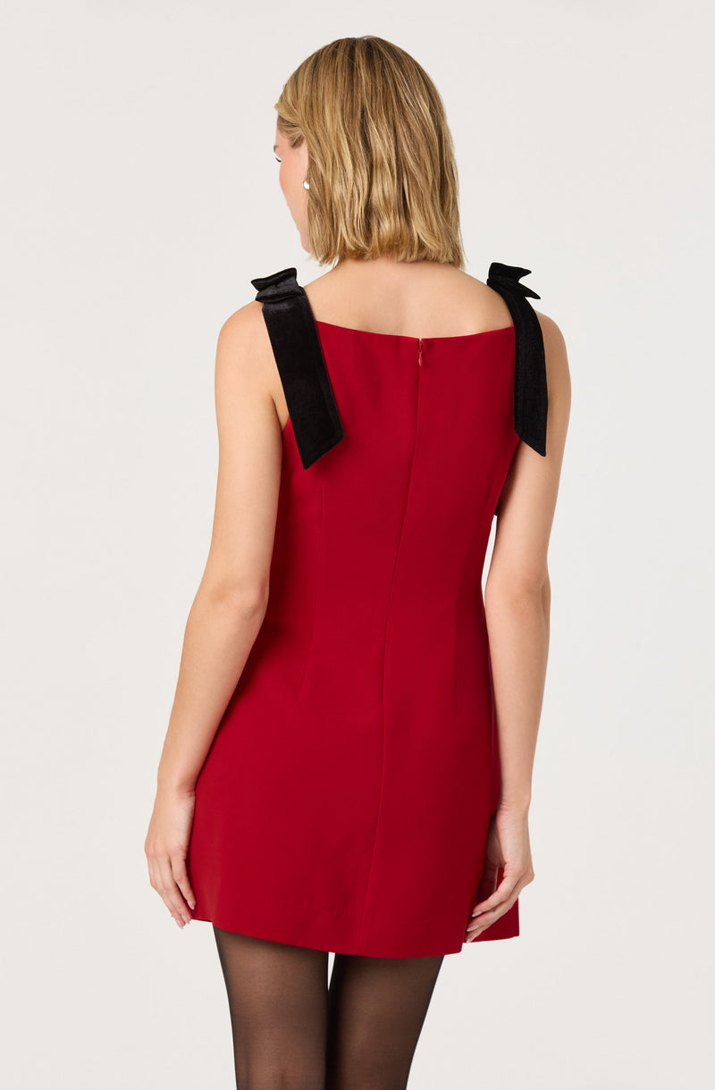 A woman is wearing a sleeveless red dress with black shoulder straps tied into bows. The dress has a simple, fitted silhouette with a central back zipper. She pairs the dress with sheer black tights.