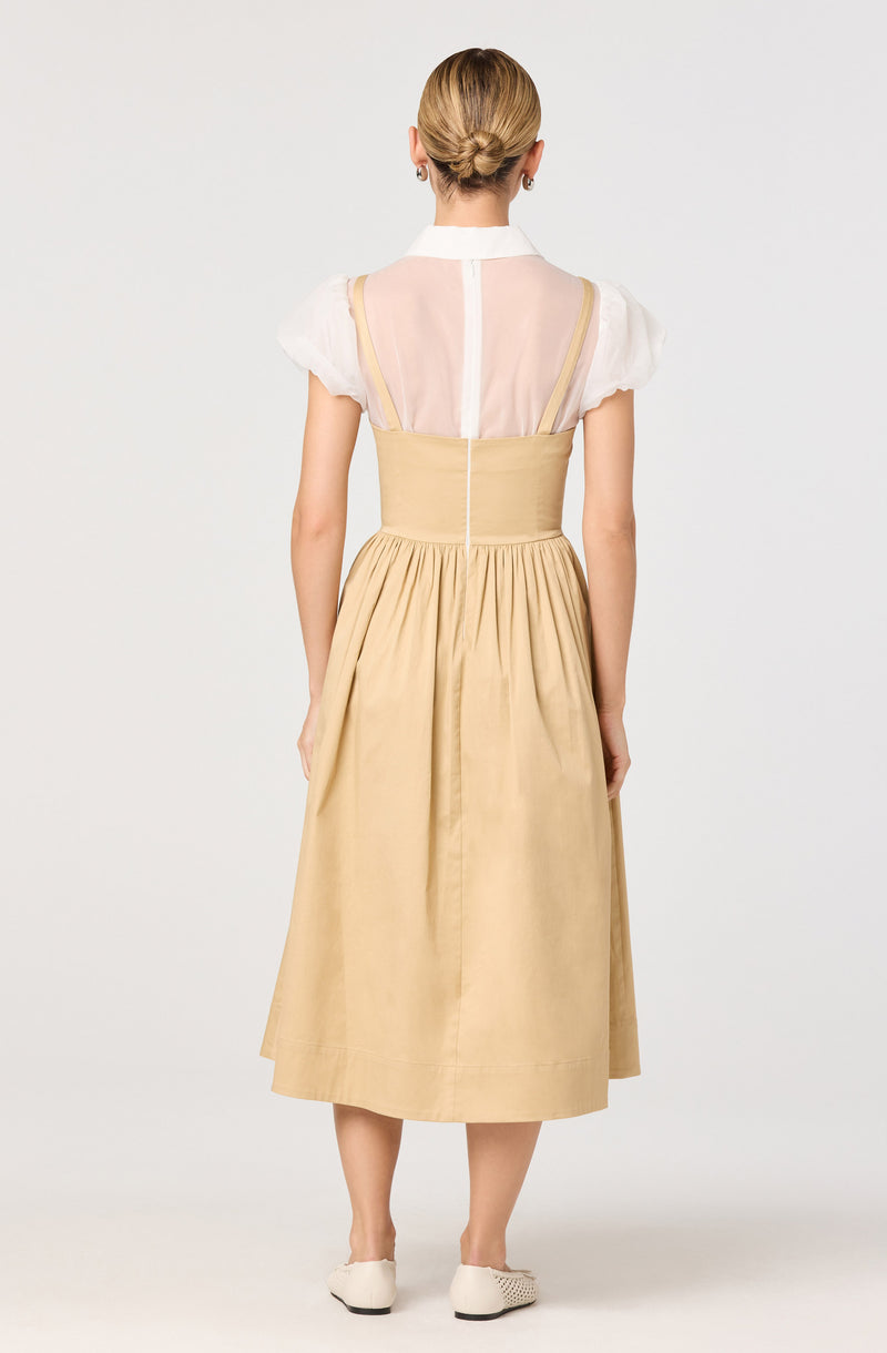 A woman is wearing a white short-sleeved blouse with puffed sleeves and a high collar. Over the blouse, she has a beige, fitted, high-waisted skirt that extends to mid-calf, with pleats at the waist. The back of the blouse is sheer, revealing shoulder straps, and the skirt is made of a lightweight, flowing fabric. She is also wearing off-white slip-on shoes with a textured design.