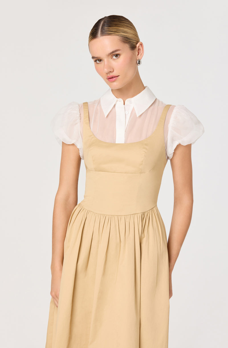 A woman is wearing a light beige sleeveless dress with a fitted bodice and a full, gathered skirt. Underneath the dress, she has on a sheer white blouse with puffy short sleeves, a pointed collar, and a button-down front. She also has on silver hoop earrings and has her hair styled in a sleek, center-parted manner.