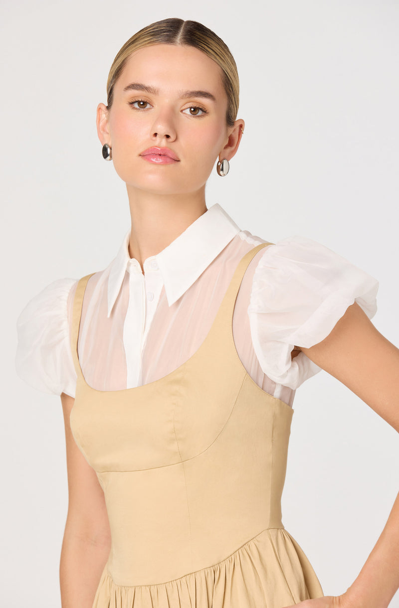 A woman is wearing a white collared shirt with puffy short sleeves layered underneath a beige sleeveless dress. The dress has a fitted bodice with a gathered skirt at the waist. She is accessorized with hoop earrings.