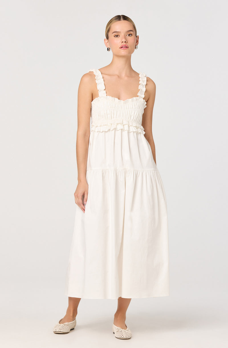 A woman is wearing a sleeveless, ankle-length dress in a light, off-white color. The dress features wide shoulder straps with ruffle details and a smocked bodice with gathered fabric. The skirt is gathered at the waist, creating a flowing, voluminous silhouette. She is also wearing a pair of white, woven or perforated flat shoes.