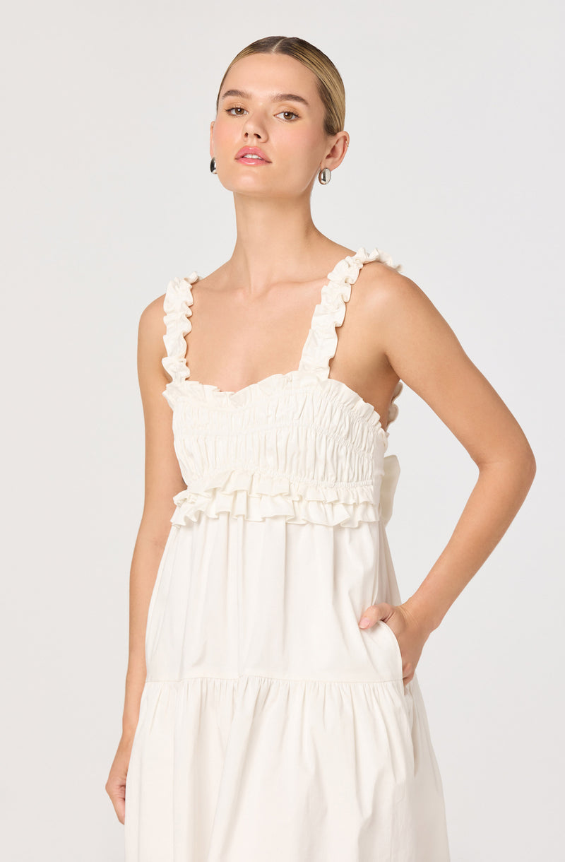 A woman is wearing a sleeveless white dress with ruffled straps and a ruffled elasticized bodice. The dress has a relaxed, flowy silhouette with a tiered skirt. She has her right hand in her pocket and is standing against a plain light background.