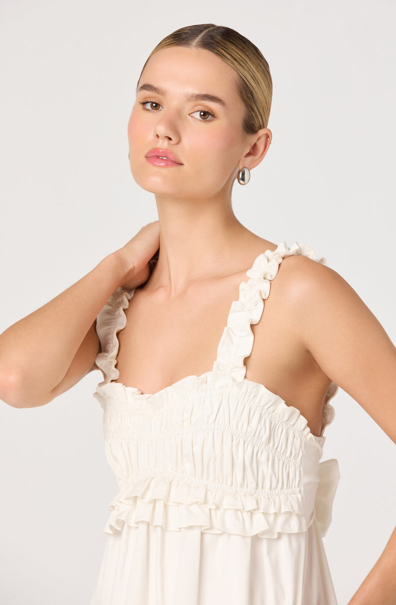 A young woman is wearing a sleeveless, cream-colored dress with ruffled straps and a gathered, ruched bodice. The dress has a flowing, lightweight fabric with ruffle details along the chest and waist. She has her hair styled straight and sleek, and she is accessorizing with small hoop earrings.