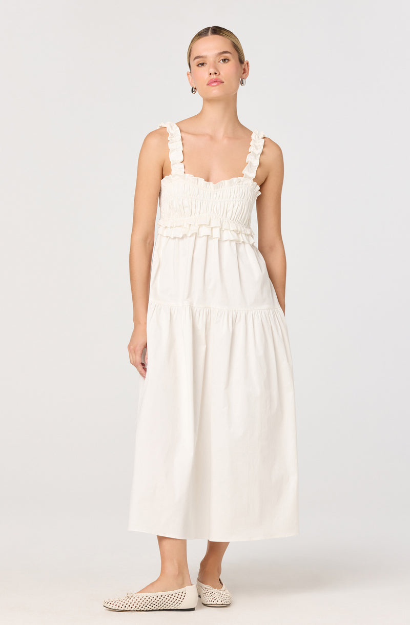 A woman is wearing a long, sleeveless, white dress with ruffled shoulder straps. The top part of the dress features a smocked, fitted bodice with ruffle details, and the skirt is loose and flowing, reaching mid-calf length. She is also wearing a pair of white, perforated flats.