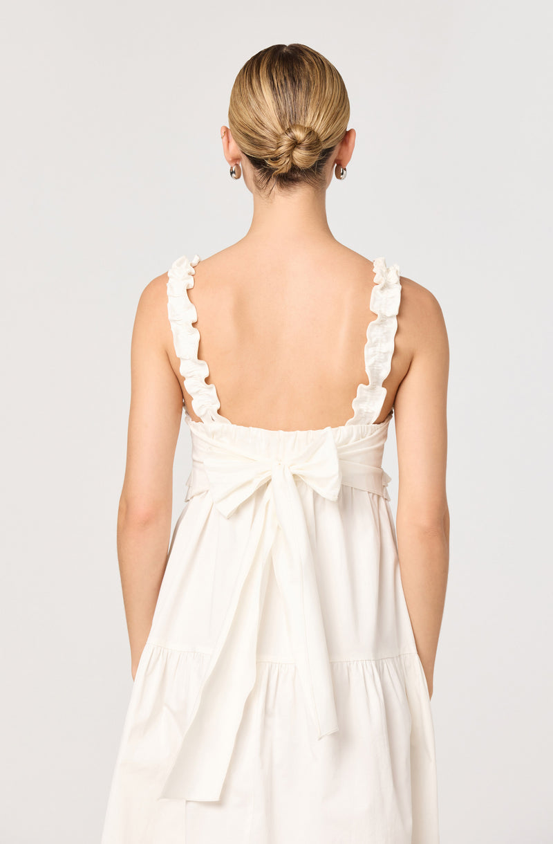 A woman is wearing a sleeveless, cream-colored dress with ruffled straps. The dress features a large bow tied at the back at waist level. Her hair is styled in a low, elegant bun, and she has on small hoop earrings.