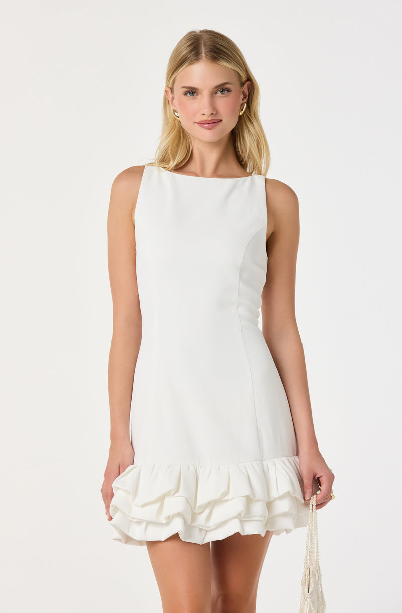 A woman is wearing a sleeveless white dress with a high neckline. The dress is fitted through the waist and hips, and features a ruffled hem at the bottom. She is also holding a small beaded purse in her right hand.