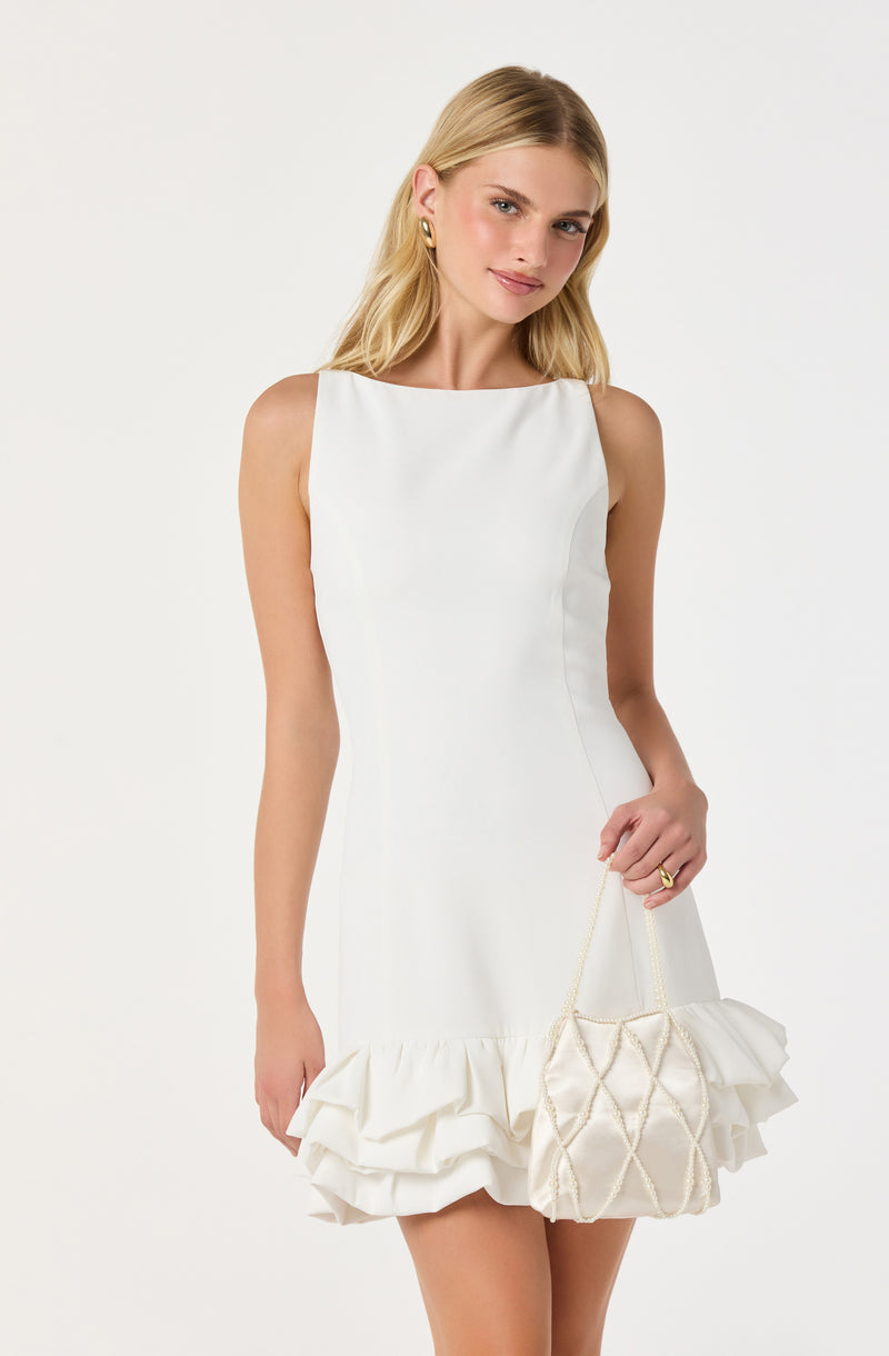 A woman is wearing a sleeveless, white dress with a fitted bodice and a ruffled hem at the bottom. She is holding a white quilted handbag with a gold chain strap.
