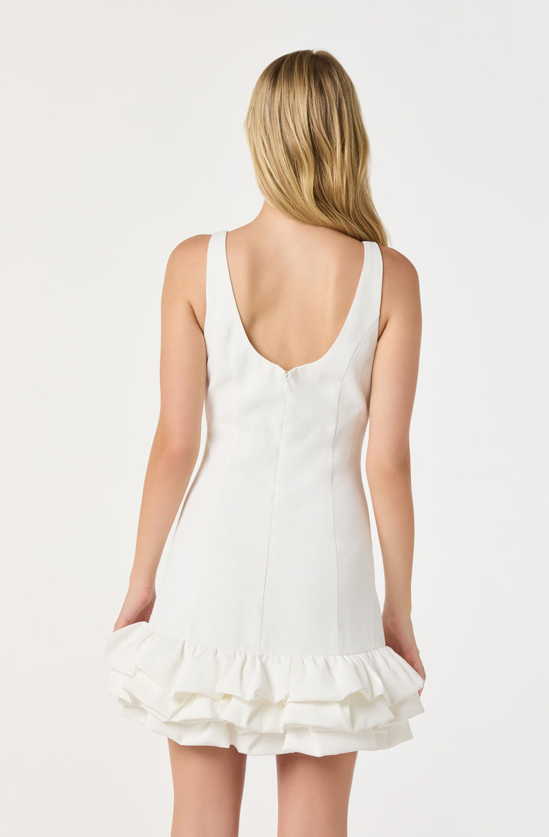 A woman is wearing a sleeveless white dress with a deep scoop back. The dress features vertical seam lines along the back and is decorated with a ruffled hem with multiple layers at the bottom.