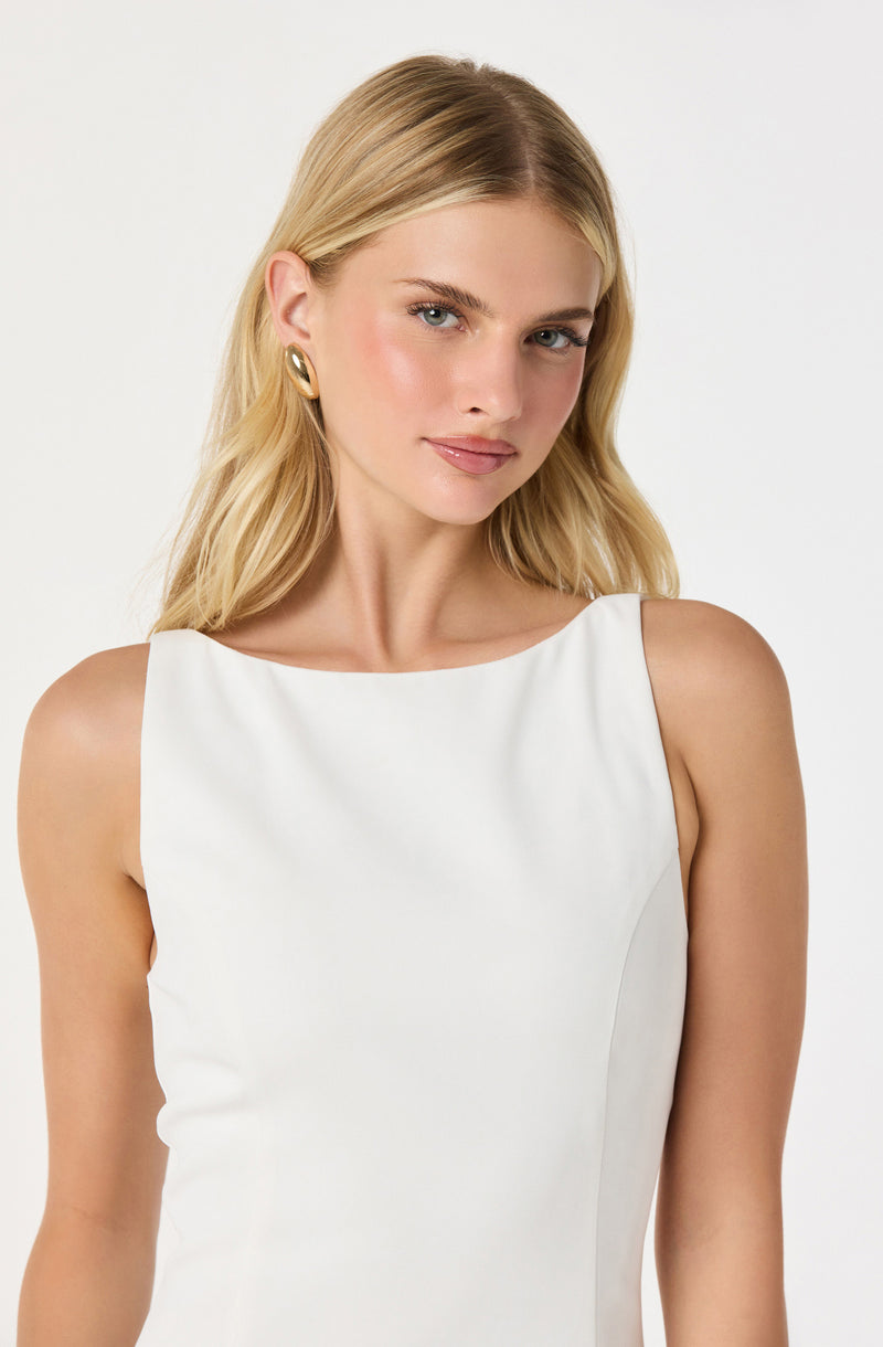 A woman is wearing a sleeveless, elegant white dress with a boat neckline. She accessorizes with large, oval-shaped gold earrings. Her blonde hair is styled in soft waves, and she has a subtle, natural makeup look.