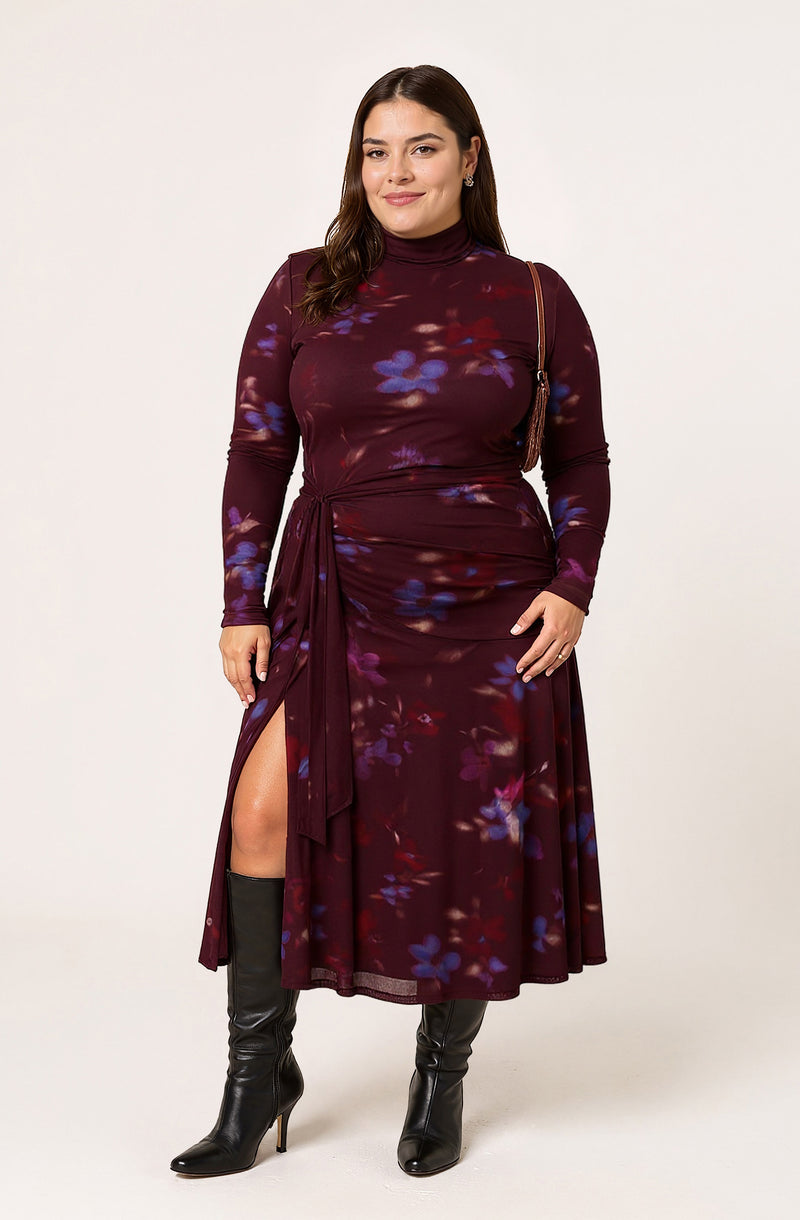 The woman is wearing a long-sleeved, high-neck dress with a dark burgundy base color and a floral pattern in shades of purple, blue, and red. The dress features a gathered waist with a side tie and a high slit on the left side. She is also wearing tall black heeled boots and has a small shoulder bag with a thin strap.