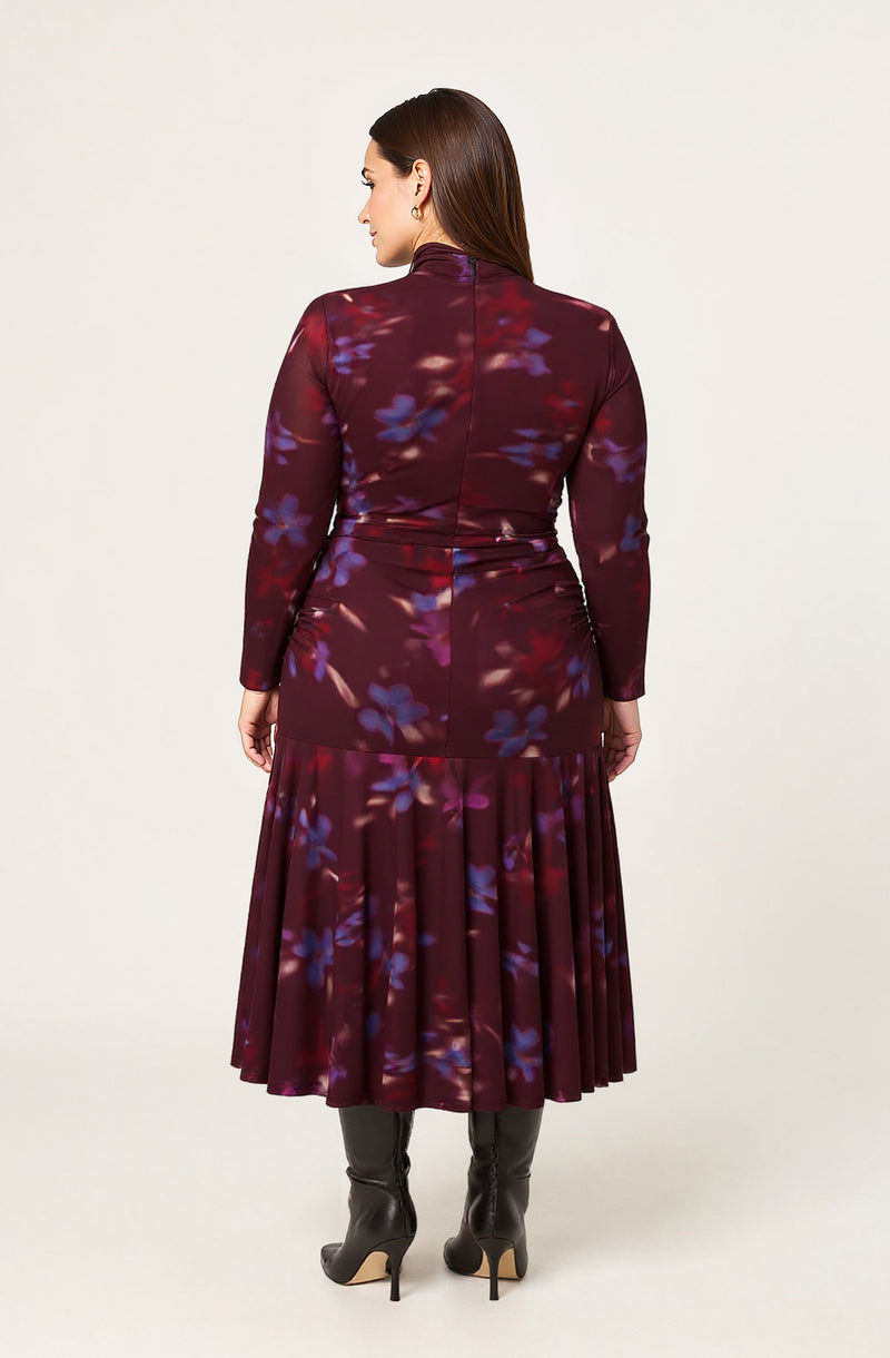 A woman is wearing a long-sleeved, fitted dress with a high neckline. The dress features a dark, rich burgundy base color with a purple and blue floral pattern. The fabric is smooth and slightly shiny. The dress is tight at the waist and transitions into a pleated skirt that falls below the knees. The woman is also wearing black thigh-high boots with high heels.