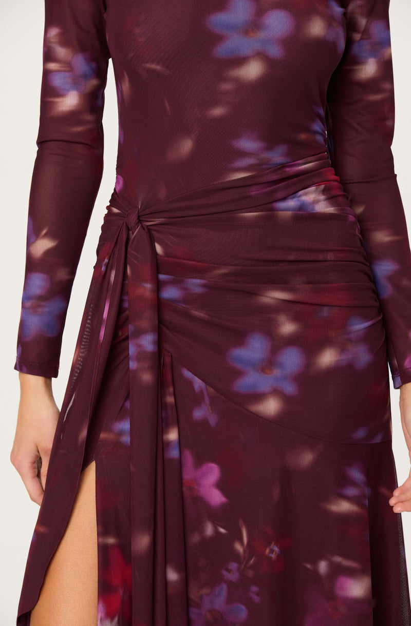 A woman is wearing a long-sleeved, fitted dress with a high slit on the left side. The dress has a deep burgundy color with a floral pattern featuring purple and pink flowers. The dress is ruched at the waist and has a tie detail on the left side, creating a draped effect.