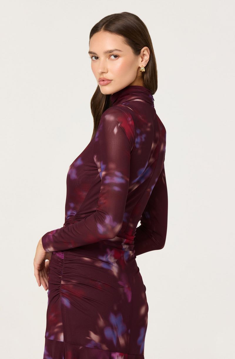 A woman is wearing a long-sleeved, fitted dress that features a high neckline and a tie-dye pattern in shades of burgundy, purple, and hints of blue. The dress has ribbed detailing around the waist area, creating a ruched effect.