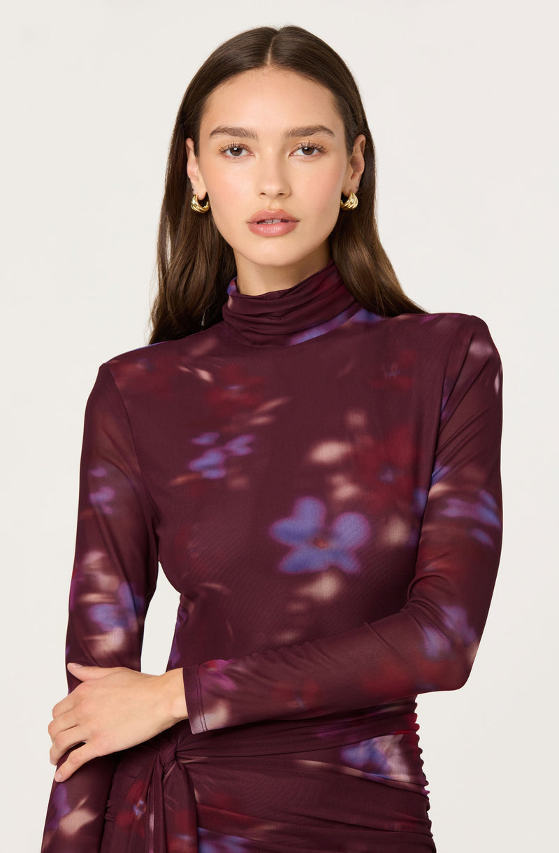 A woman is wearing a long-sleeved, fitted top with a high turtleneck. The top has a dark burgundy base color with a tie-dye pattern in shades of purple and pink. The fabric appears to be smooth and slightly shiny.