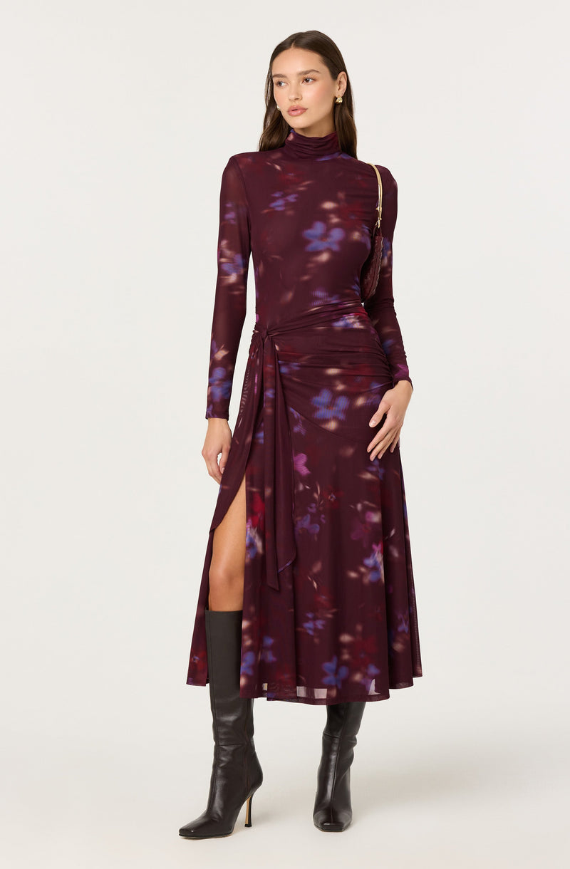 A woman is wearing a long-sleeved, high-neck, floor-length dress with a deep burgundy background and a purple and blue floral pattern. The dress features a gathered detail at the waist with a tied sash on the left side, creating a slit that reveals her left leg. She is also wearing black knee-high boots with pointed toes and stiletto heels.