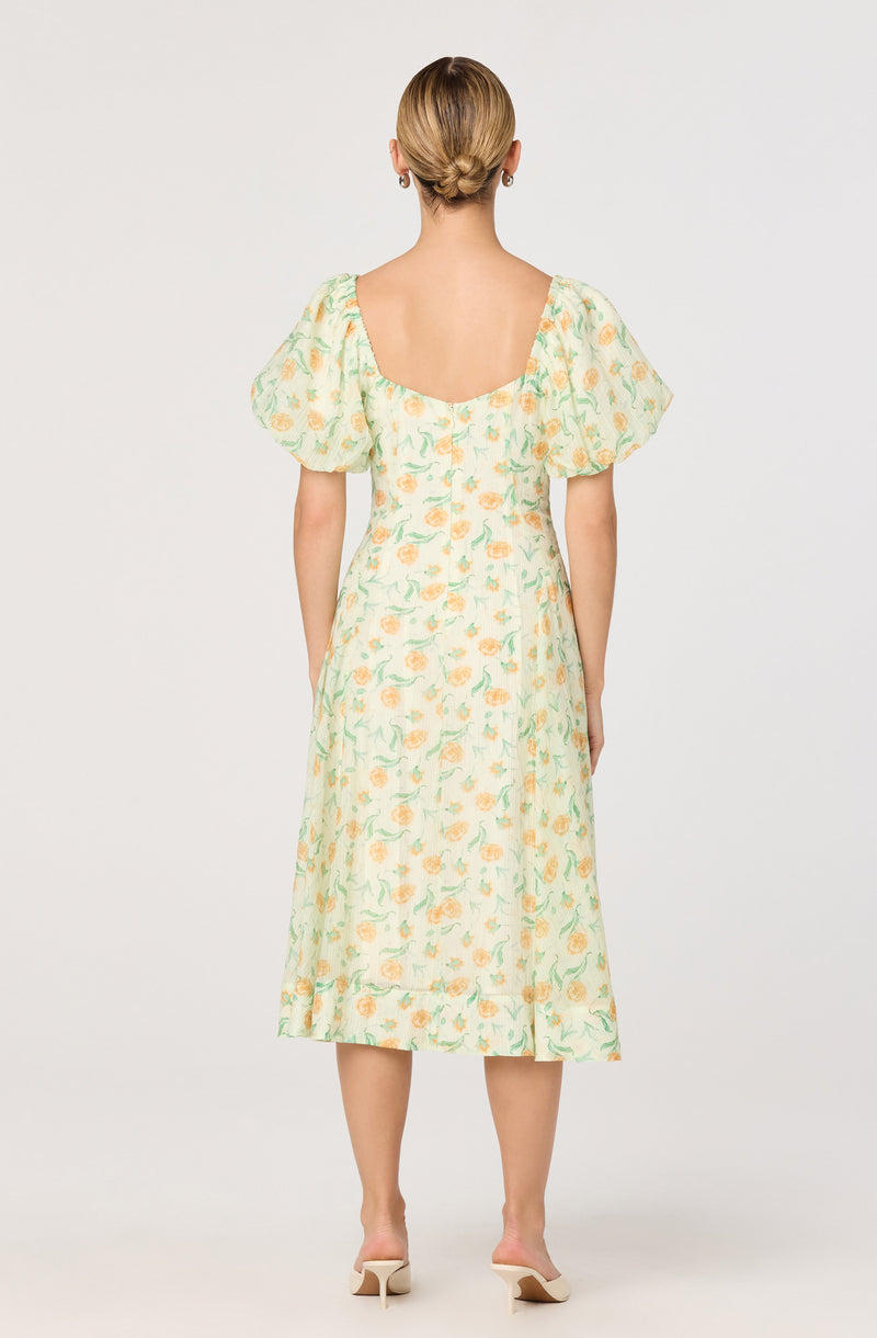 A woman is wearing a midi-length dress with a soft, pastel floral pattern featuring yellow and green colors. The dress has short, puffed sleeves and a wide neckline that extends to the shoulders, with a fitted bodice and a flared skirt. The hemline of the dress has a ruffled trim.