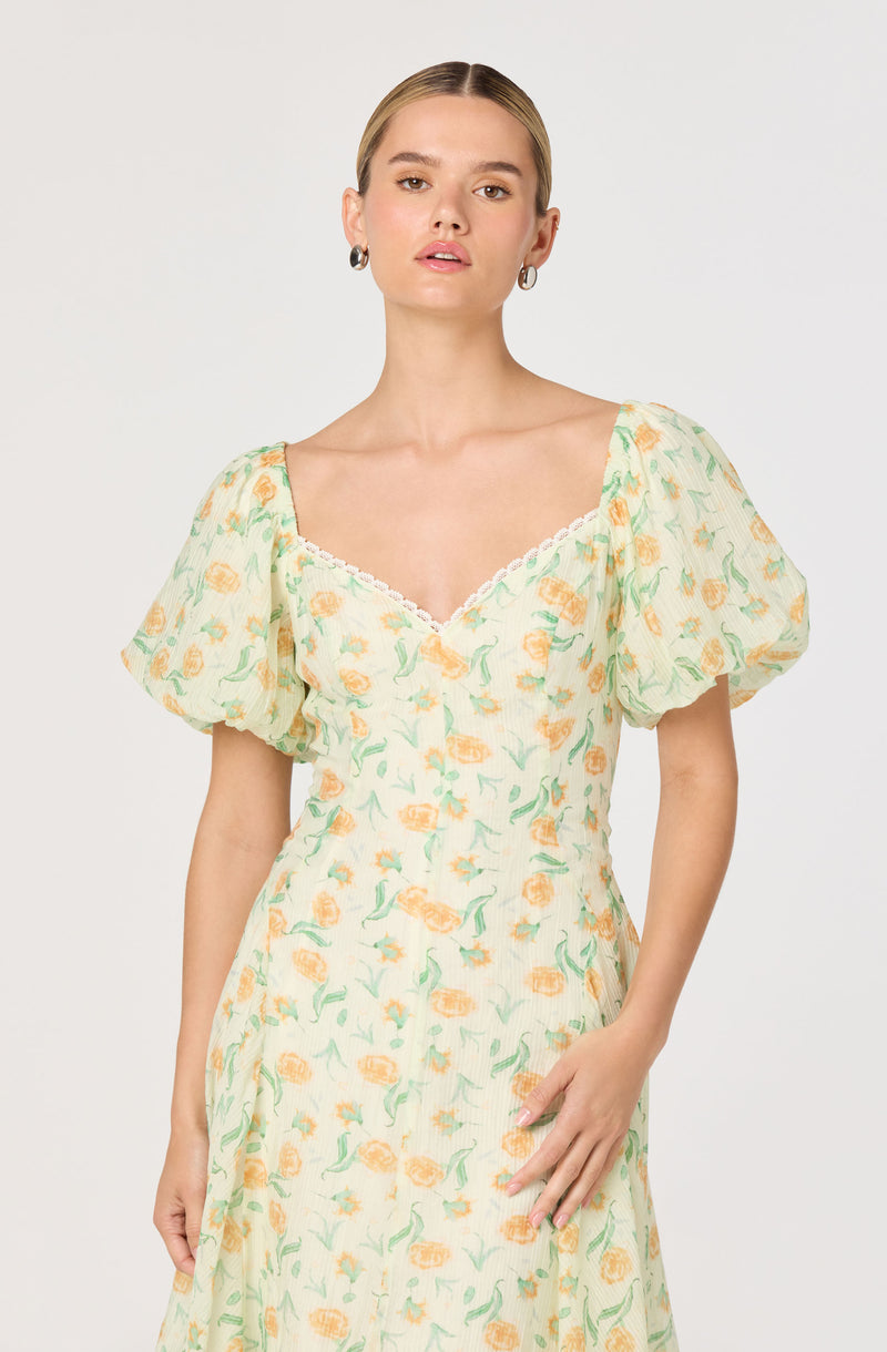A woman is wearing a light-colored dress with a floral pattern featuring yellow and orange flowers with green leaves. The dress has puffy short sleeves, a sweetheart neckline with a lace trim, and a fitted waist.