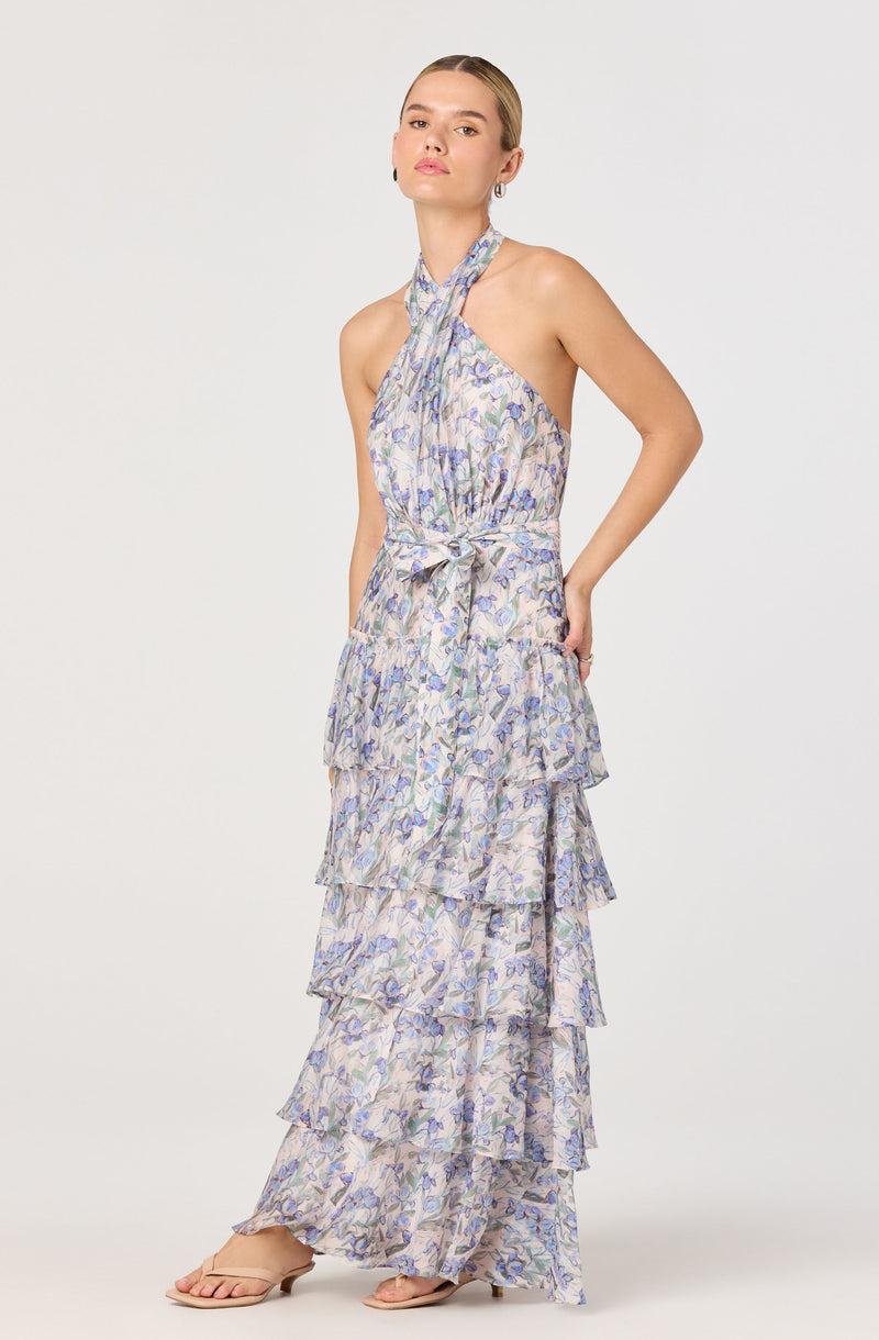 A woman is wearing a sleeveless, high-neck dress with a halter tie around the neck. The dress has a floral pattern with soft pastel colors and features multiple layered ruffles from the waist down to the hem. She is also wearing beige open-toe heeled sandals.