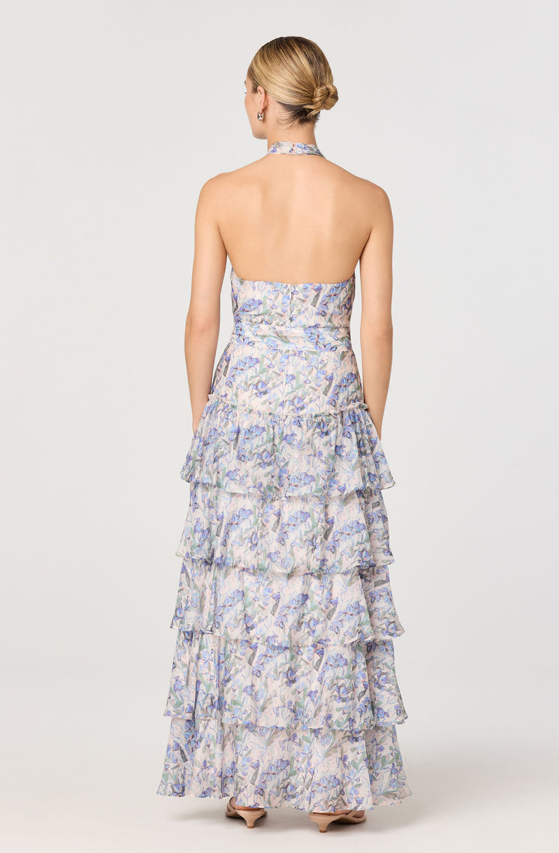 A woman is wearing a sleeveless, ankle-length dress with a halter neckline. The dress features a layered, ruffled skirt with a delicate, multicolored floral pattern on a light background.