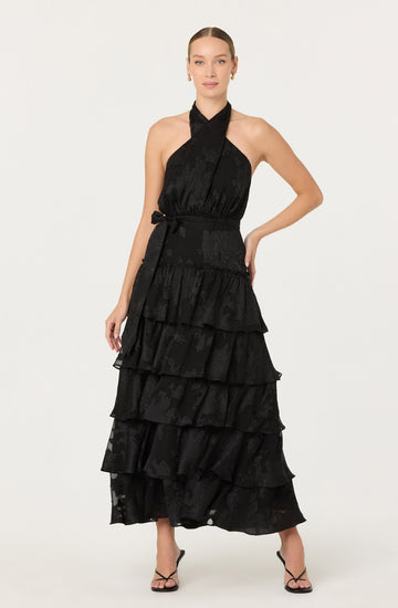 A woman is wearing a sleeveless, black halter-neck dress with a fitted waist and a tiered, ruffled skirt that reaches down to her ankles. The fabric of the dress has a subtle floral pattern. She is also wearing open-toed, high-heeled sandals with thin straps.