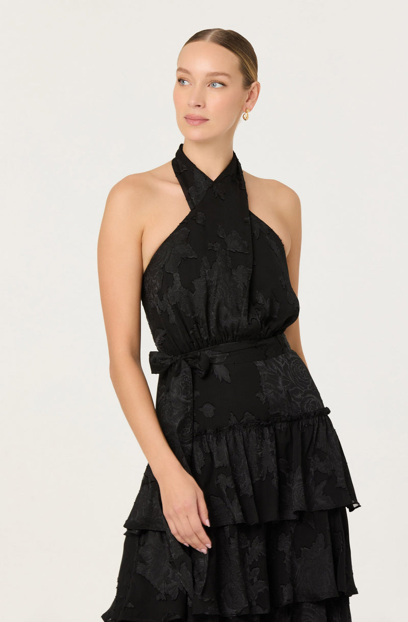 A woman is wearing a sleeveless, halter-neck dress in black with a subtle floral pattern. The dress features a waist tie at the side and tiered ruffles on the skirt. The fabric has a textured appearance with a slight sheen.