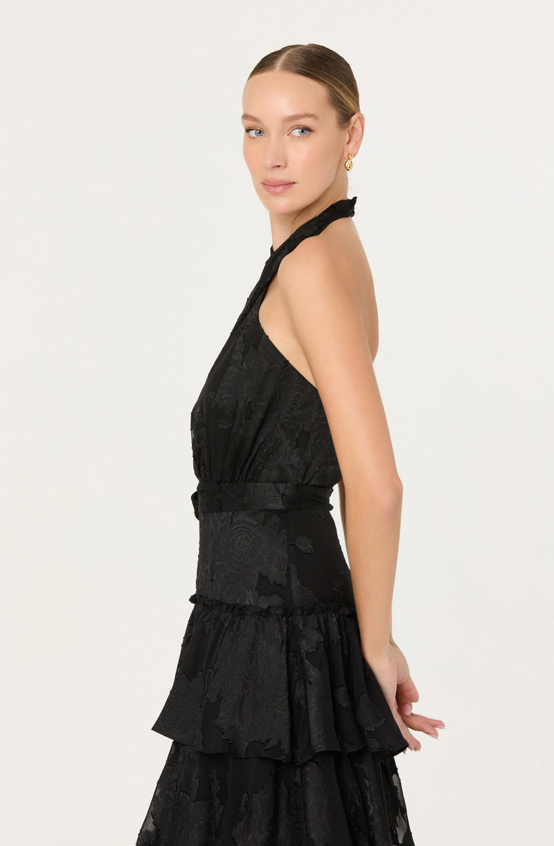 A woman is wearing a sleeveless black dress with a high neckline and a fitted waist. The dress features a textured, lace-like pattern and has a tiered, ruffled skirt.