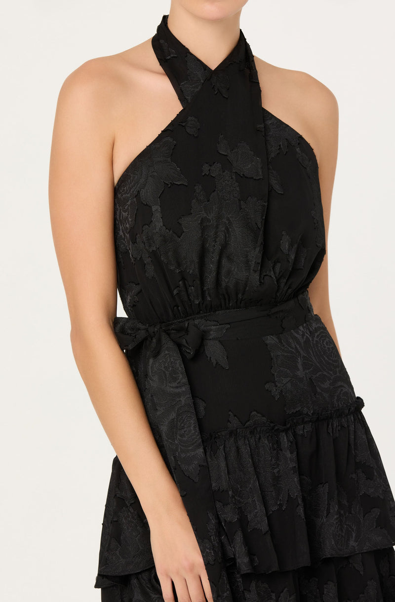 A woman is wearing a sleeveless black dress with a halter neckline that ties around the neck. The dress features a subtle floral pattern with textured floral appliqués woven into the fabric. It has a fitted waist with a matching fabric belt tied in a bow on the side, and a tiered, ruffled skirt. The overall style is elegant and sophisticated.