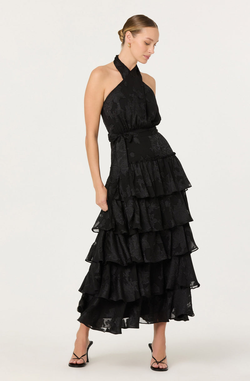 A woman is wearing a sleeveless, high-neck black dress with a fitted bodice and a full-length skirt featuring multiple layered ruffles. The fabric has a subtle floral pattern with a slight sheen. She is also wearing black open-toe high-heeled sandals.