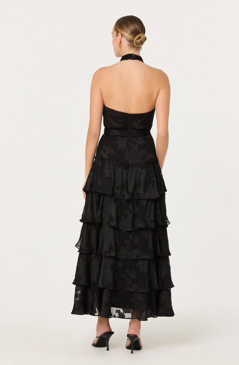 The dress is a strapless black gown with a fitted bodice and a high halter neckline. The skirt features multiple tiers of ruffled fabric, creating a layered, flowing look. The fabric has a subtle floral or jacquard pattern, adding texture to the dress. The outfit is completed with black high-heeled shoes.