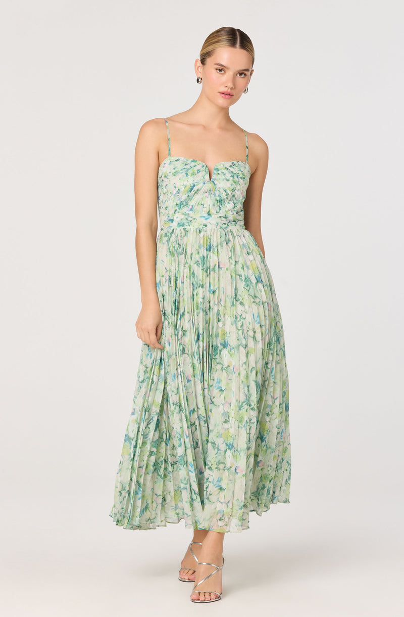 A woman is wearing a sleeveless, ankle-length dress with adjustable spaghetti straps. The dress has a fitted bodice with a sweetheart neckline and features a multicolored floral pattern in shades of green, blue, and purple on a light background. The skirt is pleated, creating a flowing silhouette. She is wearing silver strappy high heels.