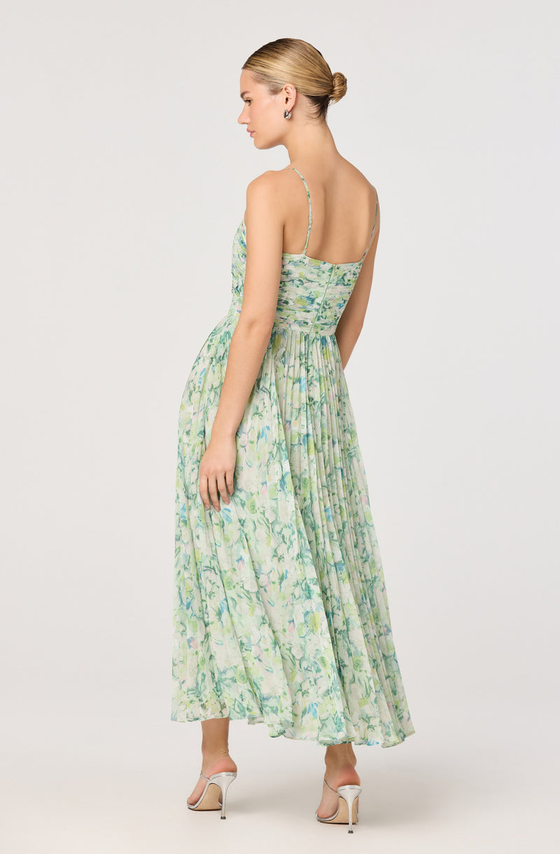 A woman is wearing a sleeveless maxi dress with thin spaghetti straps. The dress has a fitted bodice and flows down to her ankles with a pleated skirt. The fabric features a light, watercolor-like pattern with shades of green, blue, and hints of lavender and yellow. She is also wearing a pair of beige high-heeled sandals.
