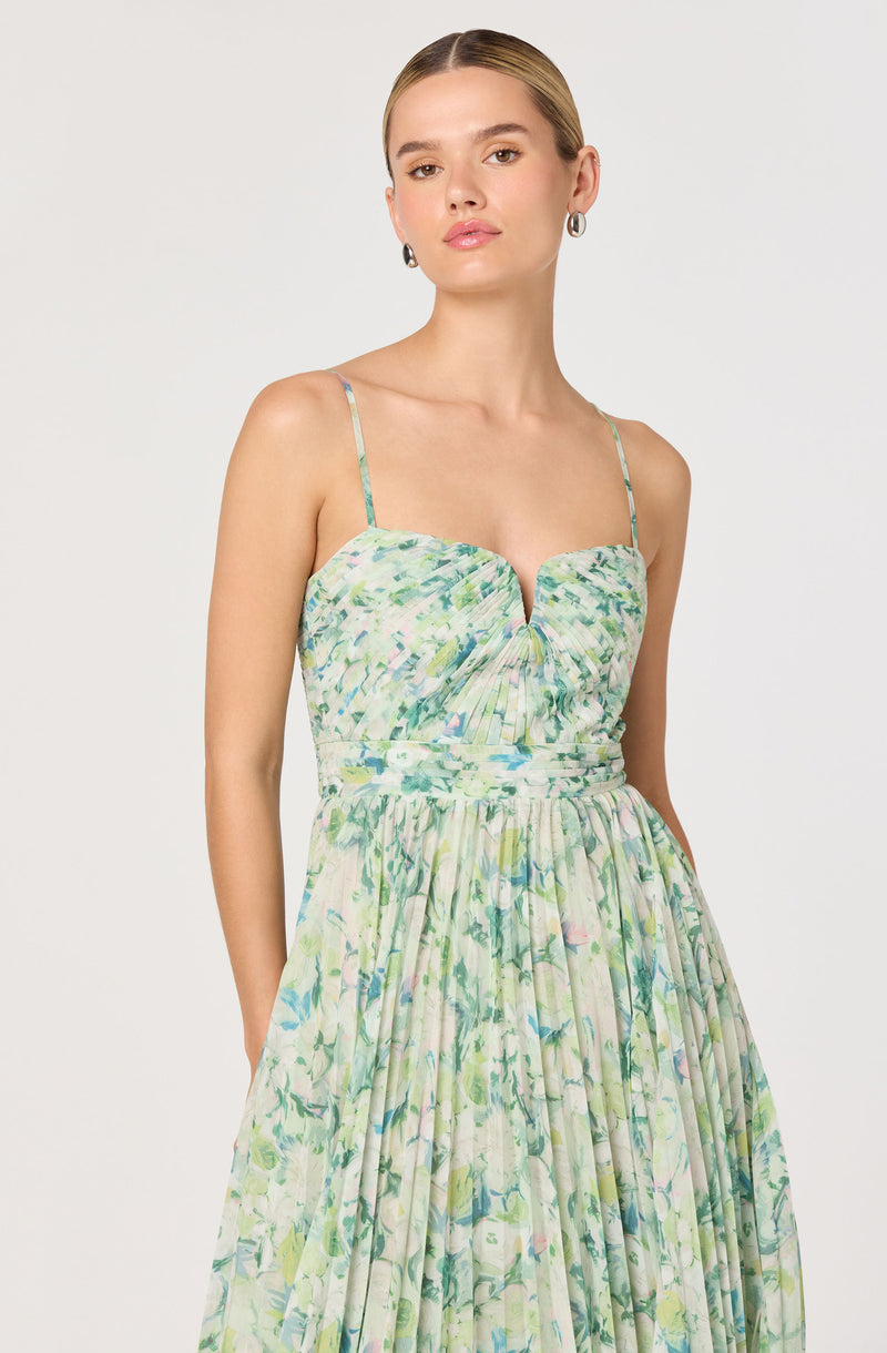 A woman is wearing a sleeveless, floral dress with thin spaghetti straps. The dress has a fitted bodice with a small V-shaped cutout at the center and features a gathered, ruched design at the bust. The skirt is full and pleated, with a light, airy fabric displaying a watercolor-like pattern of green, blue, and pink floral prints.