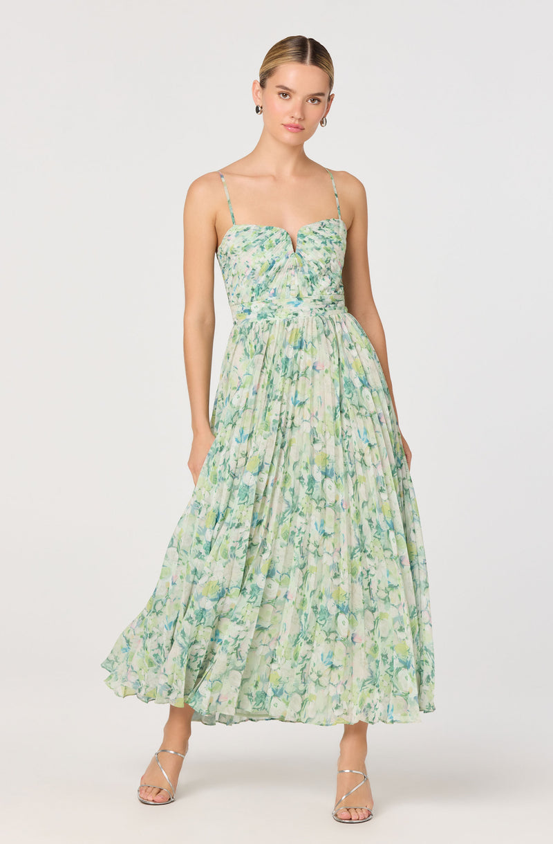The dress features a fitted bodice with a sweetheart neckline and thin spaghetti straps. The fabric has a light, airy floral pattern in shades of green, blue, and pink, with a subtle smocking detail on the top. The waistline is slightly gathered, flowing into a long, flowing maxi skirt with a soft, pleated texture. The overall look is delicate and feminine, suitable for warm weather or outdoor occasions. The model is wearing strappy heeled sandals and small hoop earrings.