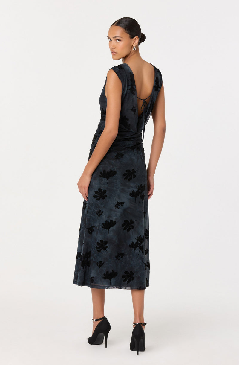 A woman is wearing a sleeveless, ankle-length dress with a dark floral pattern. The dress has a deep V back with a delicate tie detail at the top. She is also wearing high-heeled black shoes with ankle straps and small earrings.