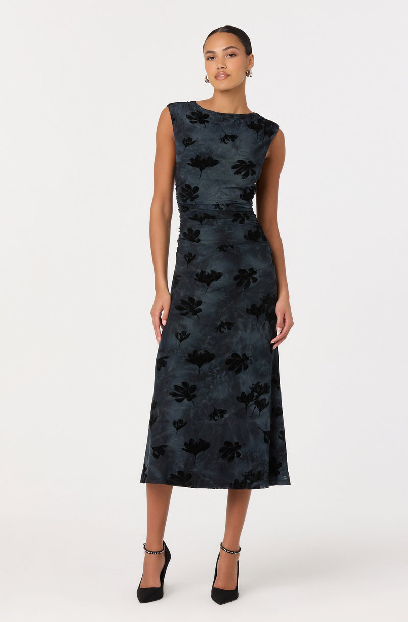 A woman is wearing a sleeveless, dark blue or black velvet dress with a floral pattern. The dress has a round neckline, a fitted waist, and a midi length that falls below the knees. She is also wearing black high-heeled shoes with ankle straps and small hoop earrings.
