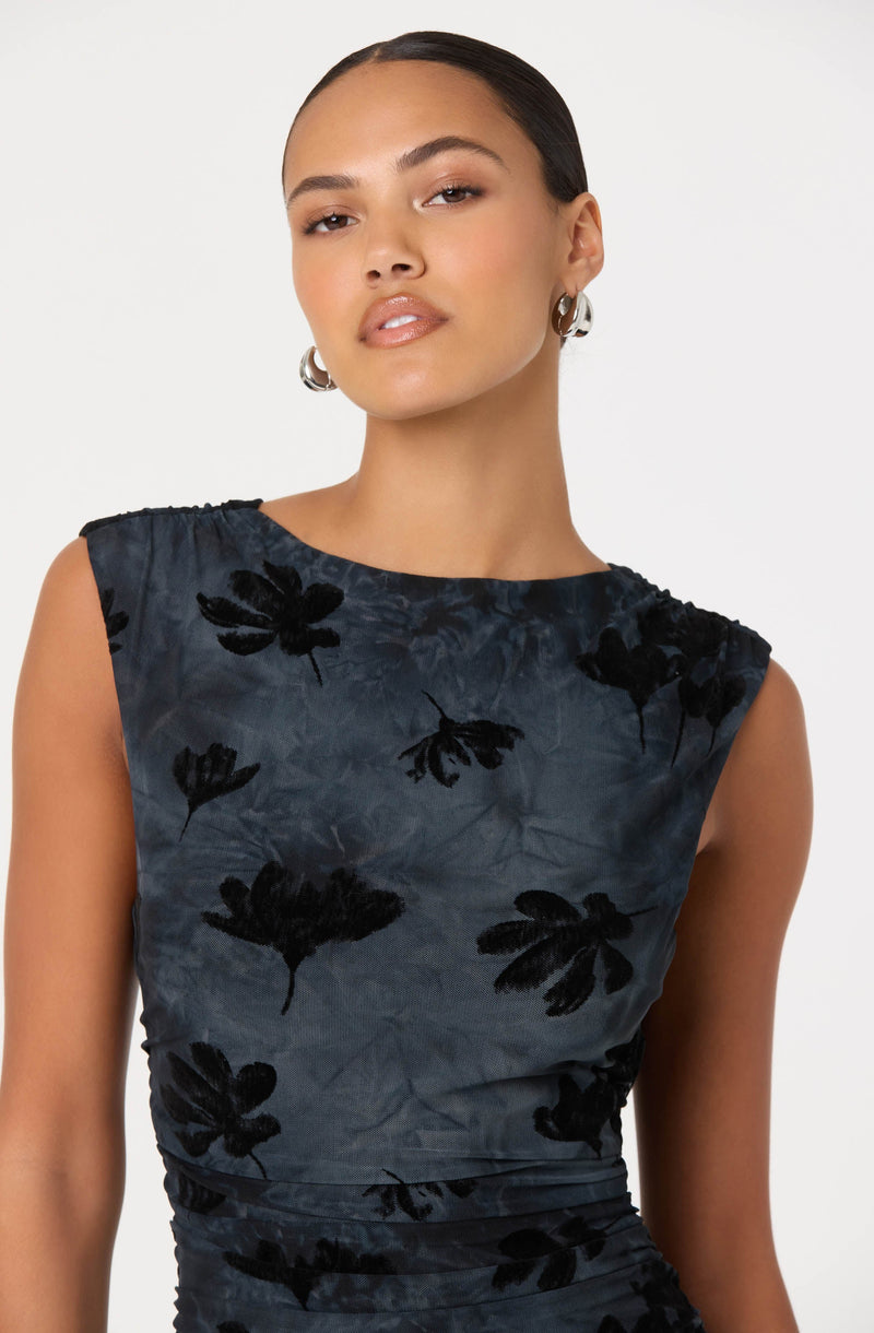 A woman is wearing a sleeveless, dark-colored dress with a round neckline. The dress features a subtle tie-dye pattern and is decorated with large, black floral motifs across the fabric.