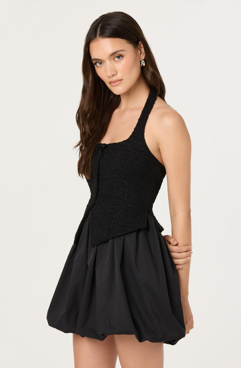A woman is wearing a sleeveless black top with a textured fabric and a fitted bodice, paired with a black bubble skirt that has a loose, voluminous shape and gathers at the waist. She is also wearing earrings with a pearl detail.