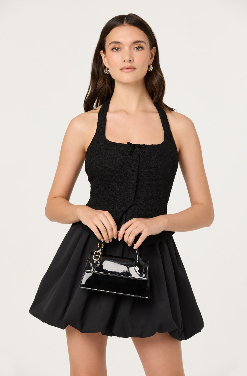 A young woman is wearing a sleeveless black textured top with a small bow at the neckline and a fitted waist. She pairs it with a high-waisted, black, puffy skirt that has a voluminous, balloon-like shape. She holds a small, shiny black handbag with a top handle and a gold clasp.