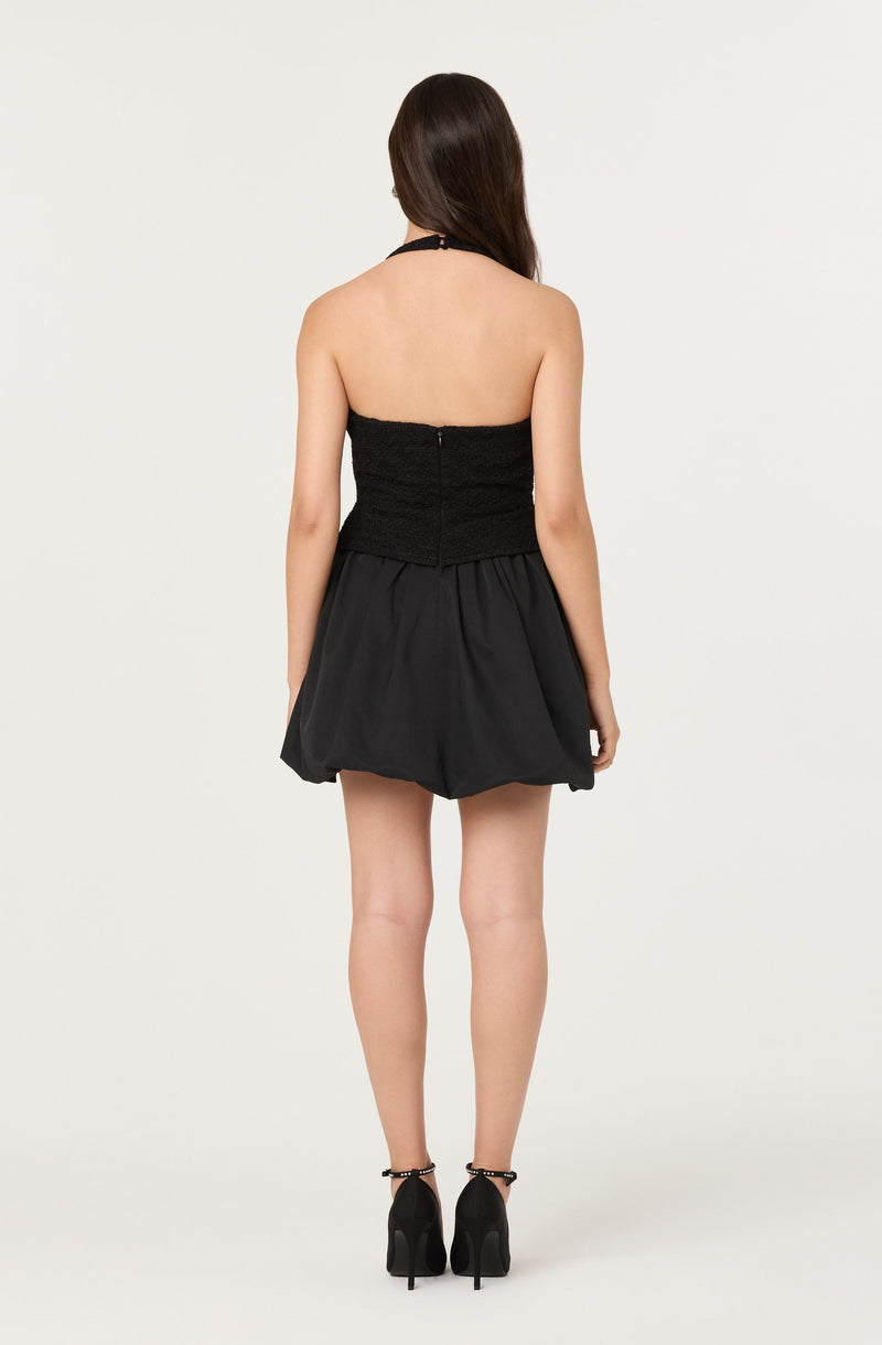 A woman is wearing a strapless black dress with a fitted bodice that has a textured or ruched appearance. The dress has a zipper at the back and a full, puffy skirt that falls above the knees. She is also wearing black high-heeled shoes with ankle straps.