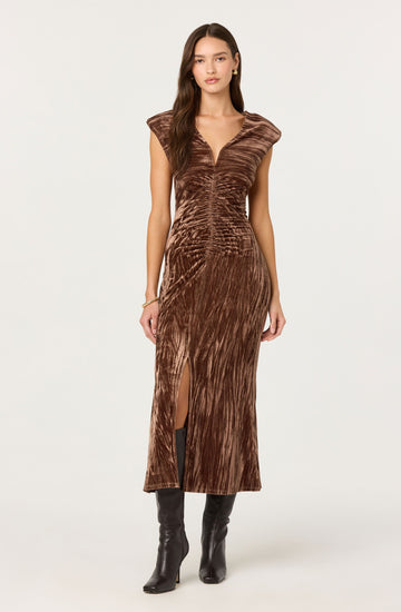 A woman is wearing a brown, velvet, sleeveless dress with a deep V neckline and shoulder pads. The dress features a gathered, ruched design at the bust and a high slit on one side, revealing her black knee-high boots.