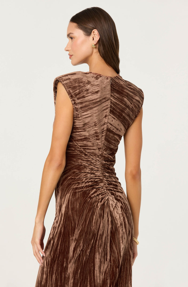 A woman is wearing a sleeveless, brown velvet dress with a textured, gathered fabric design. The dress has structured, padded shoulders and a center seam running down the back, creating a fitted silhouette. The fabric features a shiny, crinkled appearance that adds dimension to the garment.
