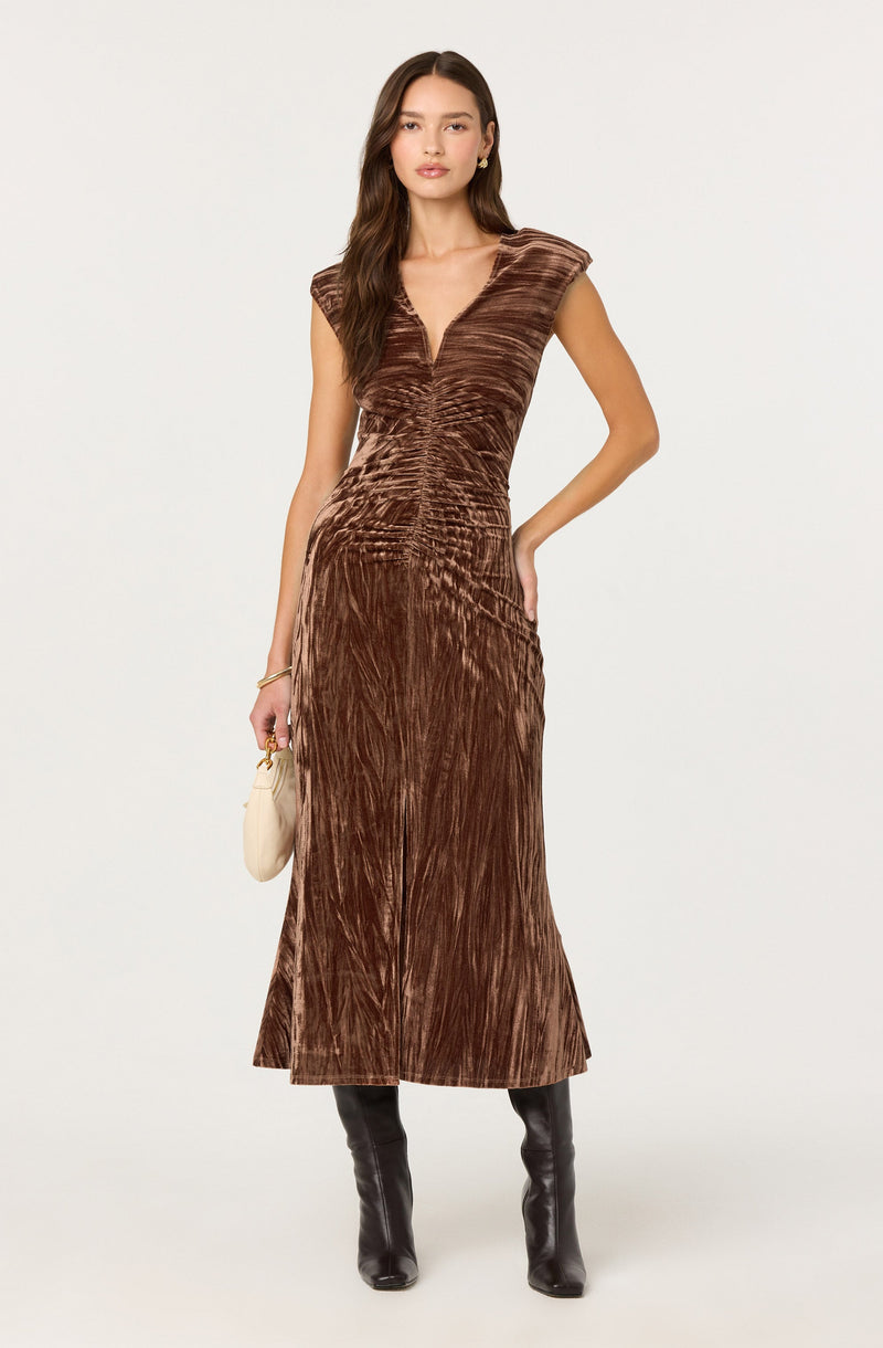 A woman is wearing a long, brown velvety dress with a V-neckline and cap sleeves. The dress has a gathered, ruched detail at the center front and features a textured, slightly shiny fabric with vertical and horizontal pleats. She is accessorizing with gold earrings, a gold bracelet, and holding a small beige handbag. She is also wearing black knee-high boots.