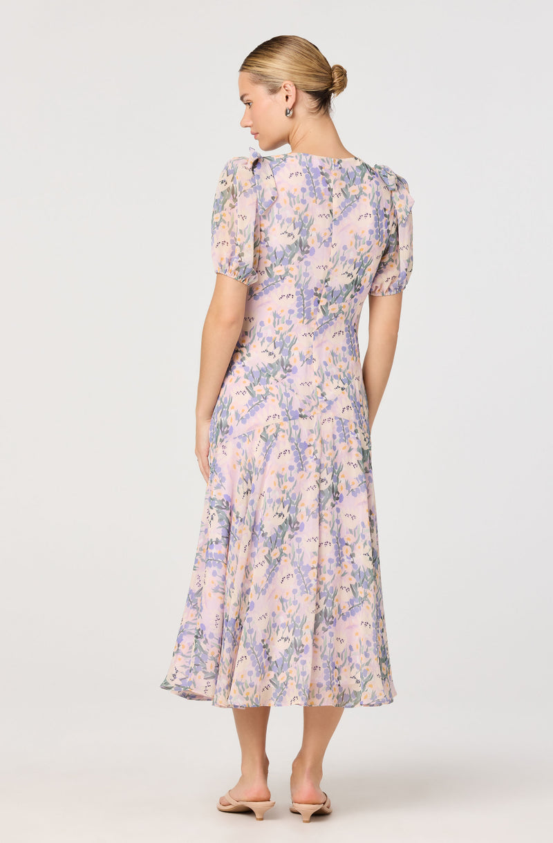 A woman is wearing a pastel-colored, floral midi dress with puffed short sleeves and a fitted waist. The dress features a light purple base with a pattern of soft pink, purple, and green floral designs. The skirt flares out slightly, creating a flowing silhouette. She is barefoot and stands against a plain, light-colored background.