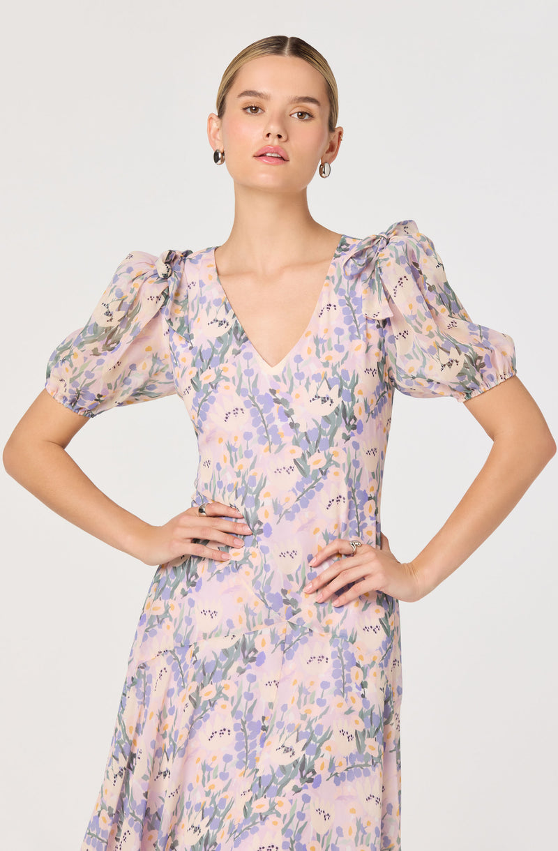 A woman is wearing a light-colored dress with a floral pattern in shades of lavender, purple, yellow, and green. The dress has a V-neckline and puffed short sleeves with tied bows at the shoulders.