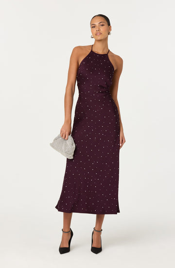 A woman is wearing a burgundy halter neck maxi dress decorated with small white polka dots. She is holding a silver mesh clutch bag and wearing black high-heeled shoes with ankle straps.