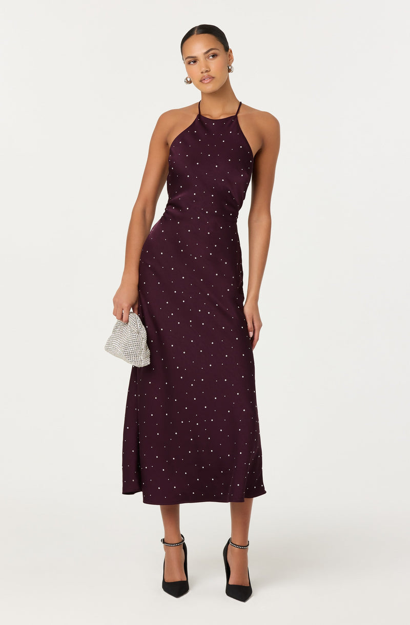 A woman is wearing a dark purple, halter-neck, ankle-length dress decorated with small white polka dots. She accessorizes with large hoop earrings, black high-heeled shoes with ankle straps, and is holding a small, metallic silver, beaded clutch purse.