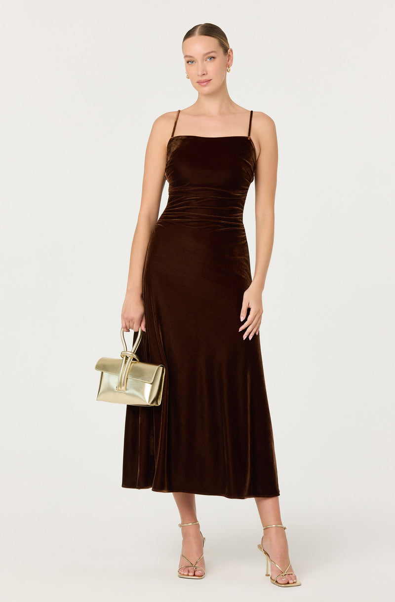 A woman is wearing a long, dark brown velvet slip dress with thin spaghetti straps. She is holding a gold metallic handbag in her right hand and is wearing high-heeled sandals with ankle straps. She has her hair pulled back in a sleek style and is standing against a plain white background.