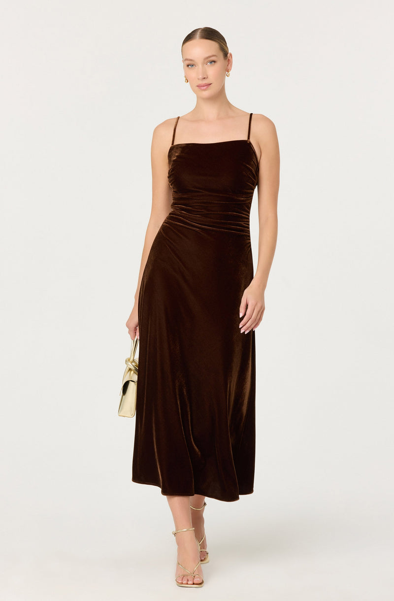 A woman is wearing a long, dark brown velvet dress with a fitted silhouette, spaghetti straps, and a straight neckline. She is holding a small, light-colored clutch purse and wearing strappy, high-heeled sandals.