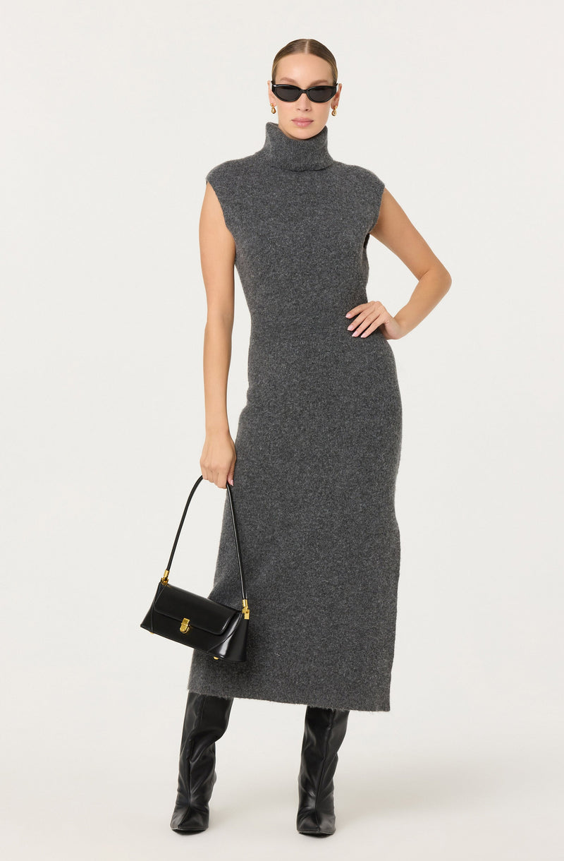 A woman is wearing a sleeveless, high-neck, long gray knit dress that extends to her ankles. She accessorizes with black sunglasses, gold earrings, and carries a small black handbag with a gold clasp. She is also wearing tall black leather boots.