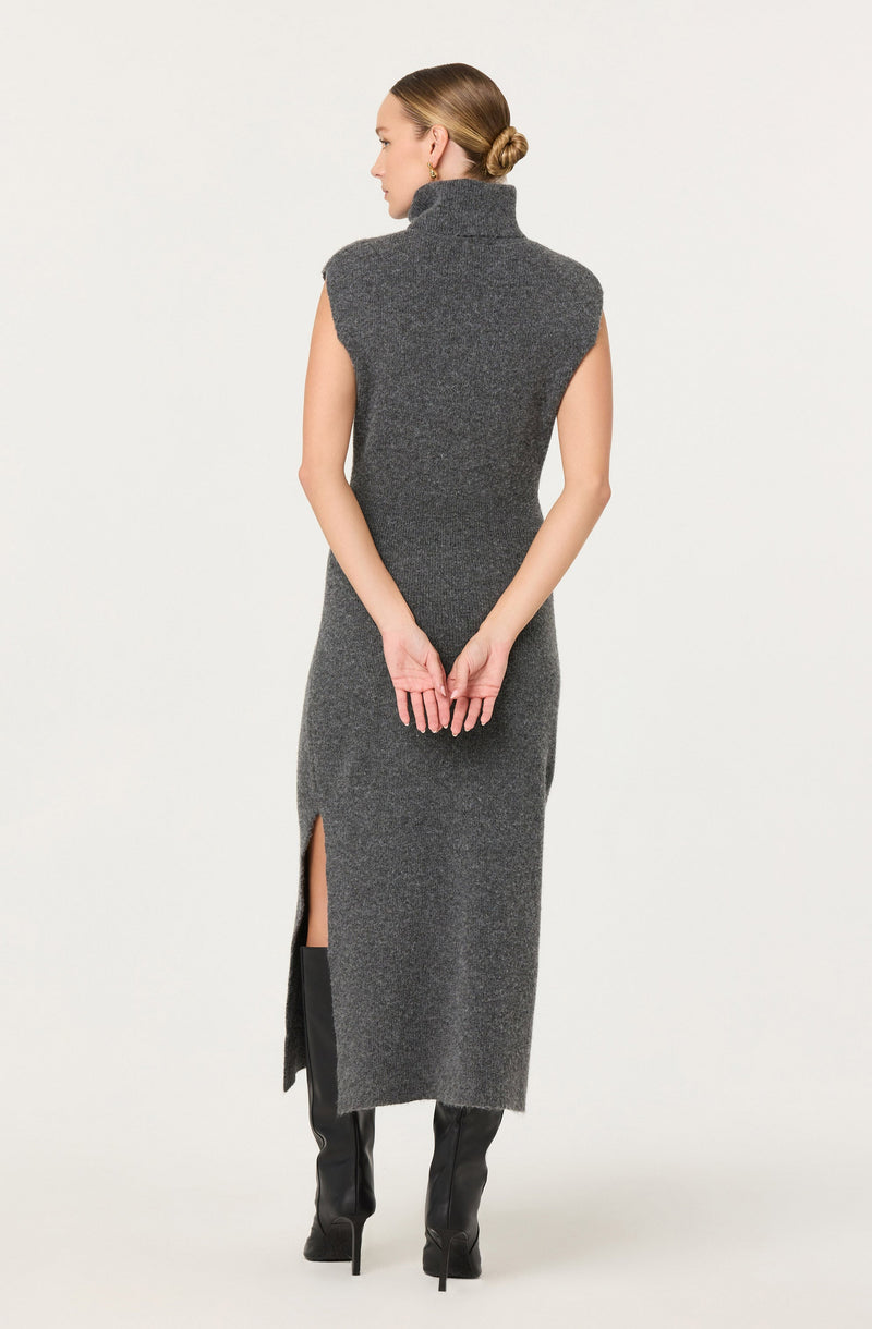 A woman is wearing a sleeveless, high-neck, gray knit dress with a side slit that reaches below the knee. The dress appears to be made of a soft, textured fabric. She is also wearing tall black boots.